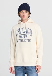 Felpa color crema con grafica "CHICAGO 1983 ATHLETIC" in blu navy; presenta una tasca frontale e cappuccio con coulisse. Abbinata a jeans blu chiaro.