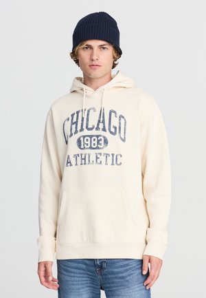 Felpa color crema con grafica "CHICAGO 1983 ATHLETIC" in blu navy; presenta una tasca frontale e cappuccio con coulisse. Abbinata a jeans blu chiaro.