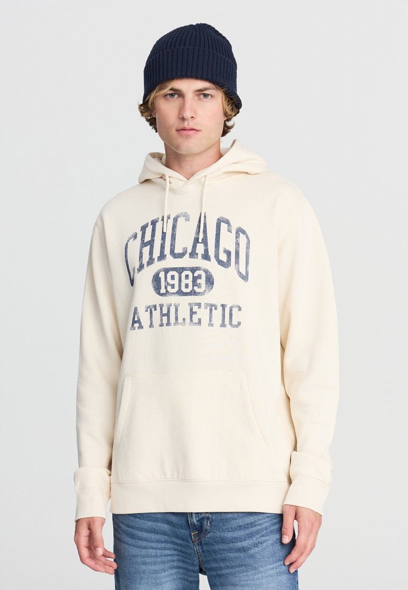Felpa color crema con grafica "CHICAGO 1983 ATHLETIC" in blu navy; presenta una tasca frontale e cappuccio con coulisse. Abbinata a jeans blu chiaro.