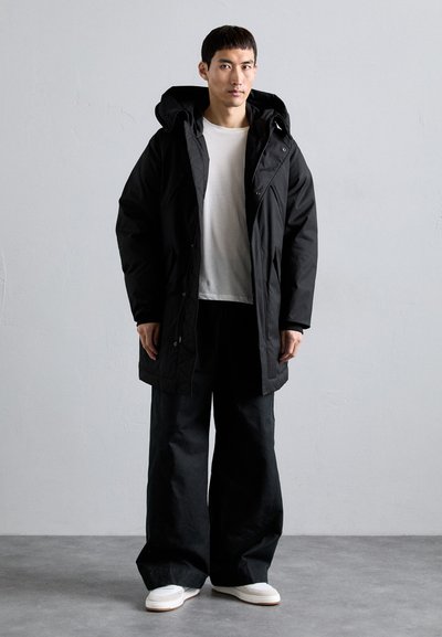Man wearing a black hooded winter coat over a white shirt, black wide-legged pants, and white sneakers, standing against a plain gray wall.