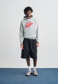 Γκρι φούτερ με κουκούλα με κόκκινο λογότυπο "Just Do It", μαύρο σορτς και ανοιχτογκρι αθλητικά παπούτσια. Ορατές λευκές κάλτσες. Απλός σχεδιασμός, casual εφαρμογή.