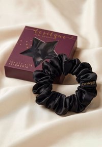 Scrunchie in seta nera con etichetta marchiata mostrata accanto a una confezione di lusso viola scuro con finestra a forma di stella su tessuto crema.
