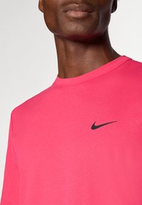 Rozā sporta t-krekls, kas izgatavots no gludas auduma, ar apaļu kakla izgriezumu un mazu melnu Nike simbolu kreisajā krūšu daļā.