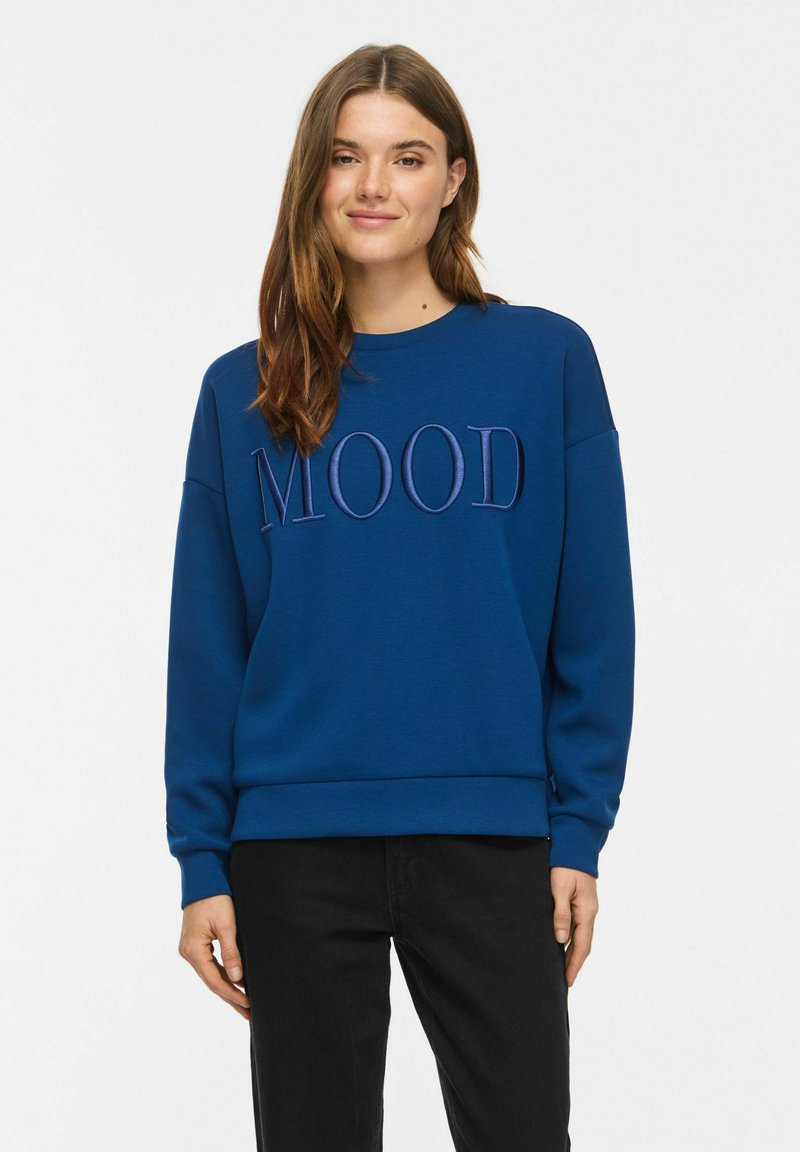Jeune femme aux longs cheveux bruns souriante, portant un sweat-shirt bleu avec le mot "MOOD" brodé et un pantalon noir, debout devant un fond blanc.
