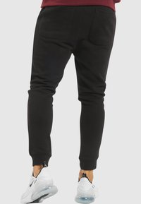 Black slim-fit joggers feature a soft, smooth texture, with an elastic waistband and tapered legs. Worn with white athletic shoes.