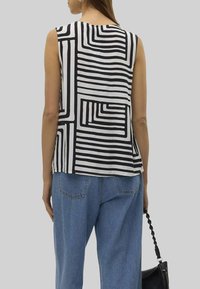 Blusa senza maniche bianca e nera, con un motivo a righe geometriche. Il tessuto appare liscio. Abbinata a pantaloni in denim larghi e una borsa nera.