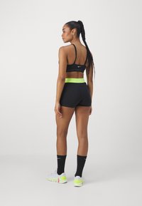 Zwarte sport-bh en shorts met felgroene tailleband. Model draagt witte schoenen met neonaccenten en zwarte sokken. Glad, aansluitend ontwerp.