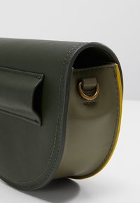 Zielono-żółta skórzana torebka na ramię o zaokrąglonych krawędziach, z widocznym przeszyciem i mosiężnym kółkiem D po boku.