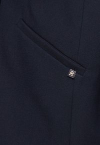 Námořnická modrá tkanina s hladkým povrchem, obsahující boční kapsu a malou zlatou visačku s logem. Design je čistý a přizpůsobený.