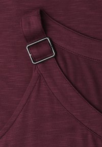 Tissu bordeaux foncé avec une boucle métallique rectangulaire sur une sangle de vêtement, montrant des coutures détaillées et une texture.