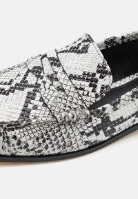 Mustvalge madussümboliga loafers, mis on varustatud tekstuuriga nahapinnaga, lõhetatud rihmaga ja siledaga musta sisemusega.