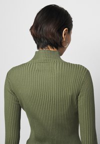 Geribbelde olijfgroene coltrui met een aansluitende silhouet, zachte textuur en hoge hals, getoond vanuit de achterkant.