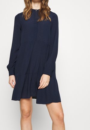 Femme portant une robe bleu marine à manches longues, longueur genoux, avec un ourlet évasé et un col à boutons, se tenant devant un fond uni.