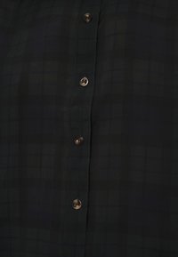 Vihreä ruudullinen nappipaitapusero, jossa on sileä rakenne. Varustettu ruskeilla napeilla ja hienovaraisella tarkkuudella kankaan pinnalla.