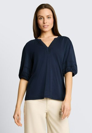 Jeune femme portant un chemisier bleu marine à manches bouffantes et un pantalon crème, debout devant un fond gris clair uni.