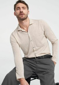 Beige langemouwen shirt van zachte stof, met een kraag, knoopsluiting en een borstzak, gecombineerd met donkergrijze broek.