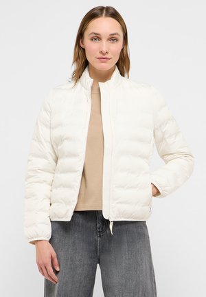 Frau trägt eine weiße Steppjacke über einem beigen Hemd und grauen Jeans, steht mit einer Hand in der Jackentasche vor einem schlichten Hintergrund.