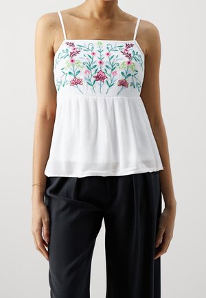 Mujer con una blusa blanca sin mangas bordada con flores multicolores y pantalones negros holgados.