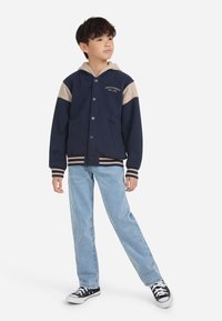 Giacca a cappuccio blu navy e beige con chiusura a bottoni, maniche a contrasto e polsini a coste, abbinata a jeans azzurri chiari e sneaker nere.