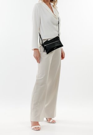 Femme portant un haut crème à manches longues, un pantalon large assorti, des talons blancs à bouts ouverts et un sac bandoulière noir avec des détails zippés.