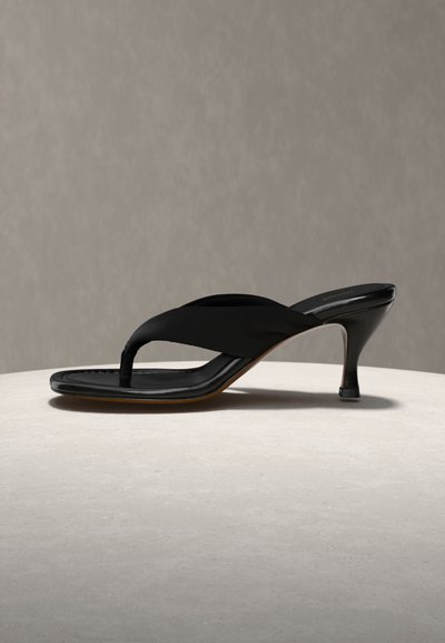 Čierna kožená ženská sandála na podpätku s priečnym remienkom a stredne vysokým ihličkovým opätkom na svetlom povrchu so sivým pozadím.