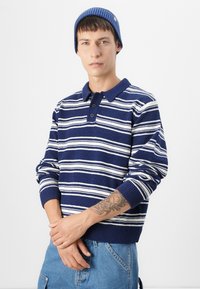 Marineblauwe en witte gestreepte pullover trui met een kraag, knoopsluiting en geribde mouwen, gecombineerd met lichtblauwe jeans.