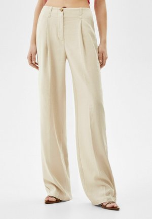 Beige wijde broek met hoge taille, voorplooien, knoopsluiting en bruine sandalen bij een staande persoon tegen een witte achtergrond.