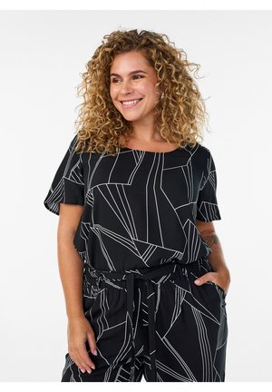 Smilende kvinde med krøllet hår iført sæt af sort top og bukser med abstrakte hvide geometriske linjemønstre og hænder i lommerne.