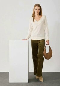 Pullover de gola em V creme, calças de veludo cotelê verde-oliva com boca de sino e botas de salto beiges. Bolsa curva castanha e suporte de exibição branco. Fundo neutro.