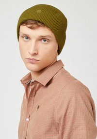 Olijfgroene gebreide beanie met ribbelstructuur en een klein logo op de voorkant. Gepaard met een lichtroze geblokt shirt.