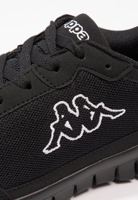 Svarta sportskor med nät- och mockamaterial; har svarta snören, texturerad sula och vitt broderat logotyp på sidan.