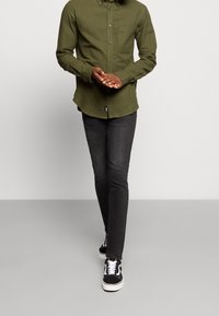 Uomo che cammina in avanti indossando una camicia verde oliva con bottoni, jeans skinny neri e sneakers bianche e nere su uno sfondo semplice.