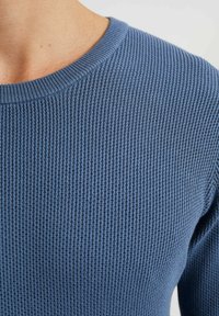 Blå strukturerad stickad tröja med rund halsringning och ribbade mudd, med en närbild som framhäver dess tygmönster och struktur.