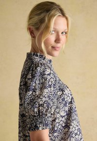 Blonda sieviete ar ziliem acīm, kura valkā zelta riņķveida auskari un tumši zilu ziedu augstu apkakles blūzi, maigi smaidot uz vienkārša gaiši pelēka fona.