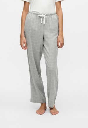 Persoon die lichtgrijze geruite broek met trekkoord en witte top draagt, staat op blote voeten tegen een effen witte achtergrond.