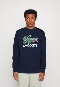 Felpa navy con logo di un coccodrillo verde e testo "LACOSTE" in bianco. Collo e polsini a coste. Tessuto morbido e strutturato.