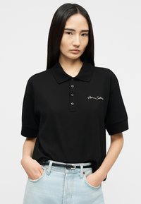 Γυναίκα με μακριά σκούρα μαλλιά που φορά μαύρο πόλο με λογότυπο, μαζεμένο μέσα σε ανοιχτόχρωμο τζιν με μαύρη ζώνη, χέρια στις τσέπες, ουδέτερο φόντο.