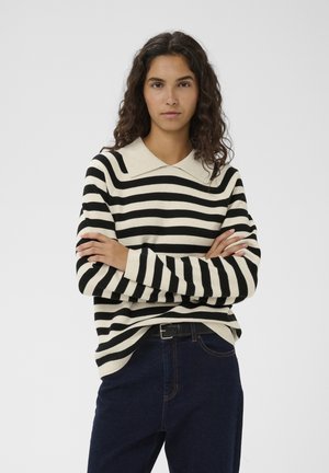 Žena s dlhými kučeravými vlasmi, oblečená v čierno-bielom pruhovanom pulóvri s golierom a tmavých džínsoch, stojaca so založenými rukami.