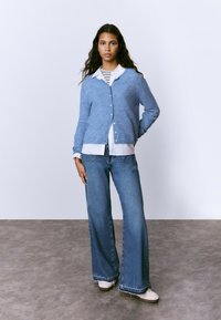 Cárdigan azul claro con botones, superpuesto a una camisa blanca y una camiseta de rayas, combinado con jeans de pierna ancha azul y zapatillas blancas.