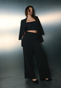 Čierne široké nohavice so sequínmi a vysokým pásom, kombinované s priliehavým čiernym crop topom a voľným čiernym blejzrom, zobrazené na kovovom pozadí.