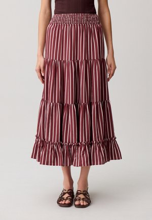 Mujer con una falda de capas color burdeos con rayas verticales blancas y sandalias marrones con tiras, de pie contra un fondo claro y liso.