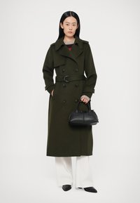 Olijfgroene wollen trenchcoat met dubbelzijdig knoopsluiting, ceintuur in de taille en grote knopen. Gecombineerd met witte, wijd uitlopende broekspijpen en een zwarte handtas.