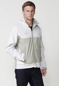 Veste légère pour homme avec une partie supérieure blanche et une partie inférieure grise clair. Caractéristiques : col haut, poches zippées et poignets élastiques.