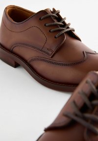 Chaussures habillées en cuir marron avec une finition lisse, présentant des coutures décoratives, un laçage et un petit talon carré.