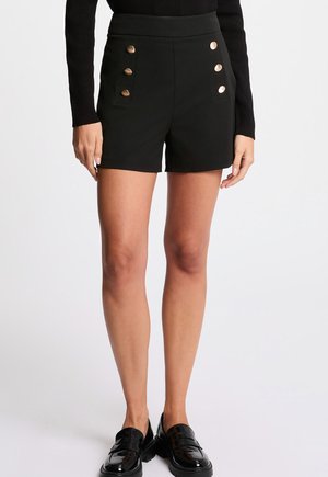 Shorts noirs taille haute en tissu lisse, avec cinq grands boutons dorés à l'avant et des poches latérales. Associés à des mocassins noirs.