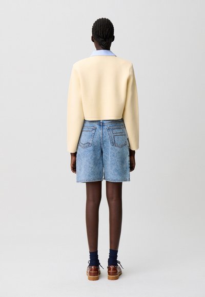 Maglione giallo tagliato, pantaloncini di denim blu, calzini scuri, scarpe marroni. Design semplice con spalle strutturate e materiali a contrasto.