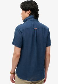 Hombre de cabello oscuro que lleva una camisa azul marino de manga corta y pantalones beige, visto desde atrás sobre un fondo blanco.