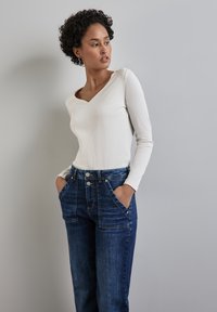 Weißes, geripptes Langarmoberteil mit V-Ausschnitt, kombiniert mit blauen Denim-Jeans mit hoher Taille, großen Fronttaschen und einer Zwei-Knopf-Verschluss.