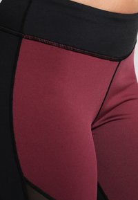 Leggings a vita alta con pannello bordeaux e accenti neri, realizzati in tessuto elasticizzato con una superficie liscia e cuciture a contrasto.