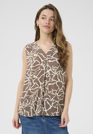 Kobieta o długich falowanych włosach, ubrana w bezrękawnik z brązowo-białym wzorem oraz niebieską spódnicę dżinsową, spoglądająca lekko na bok.